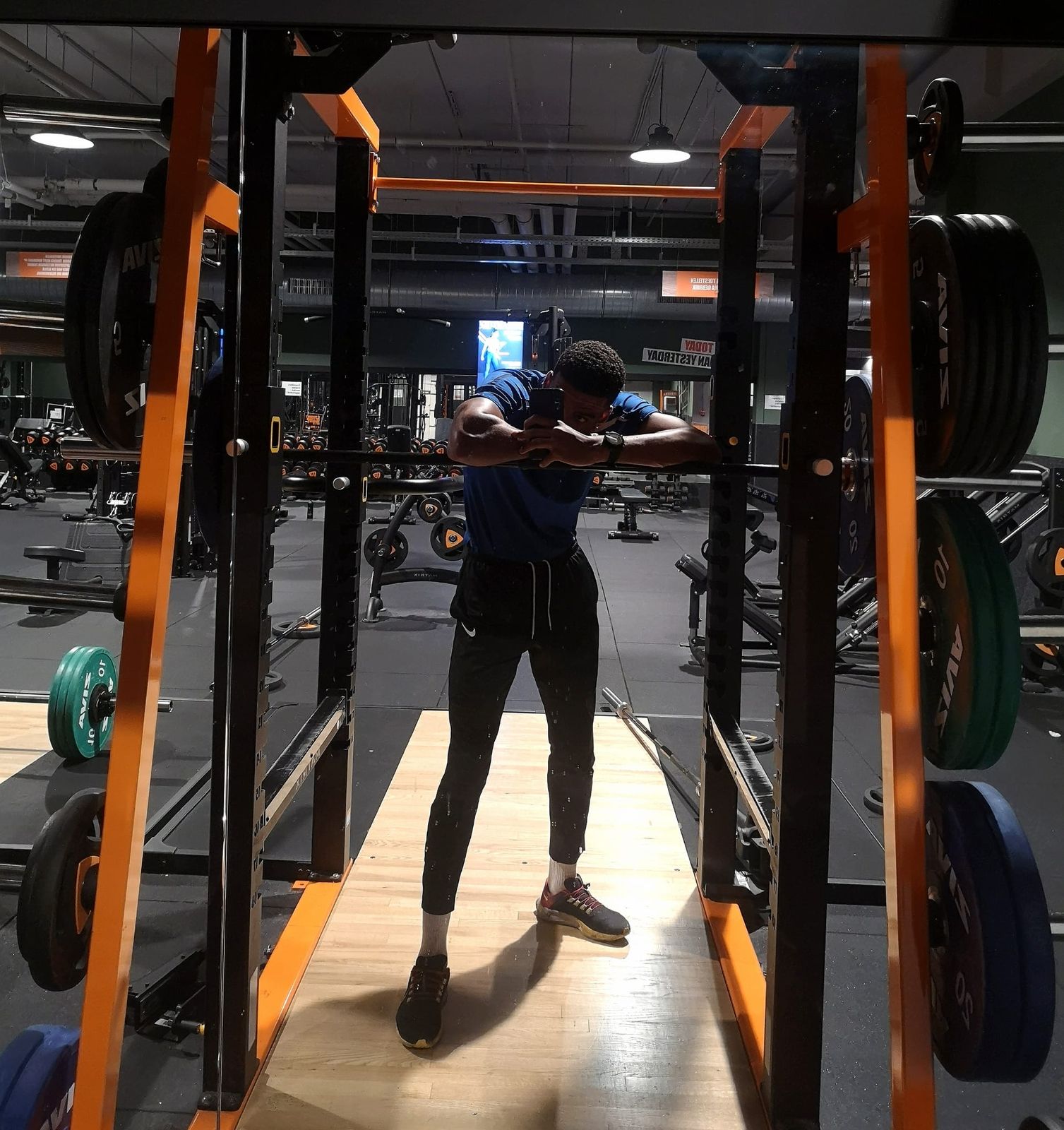 Manu in the gym, inside a rack and leaning on the bar, in a blue T-shirt with plates around him