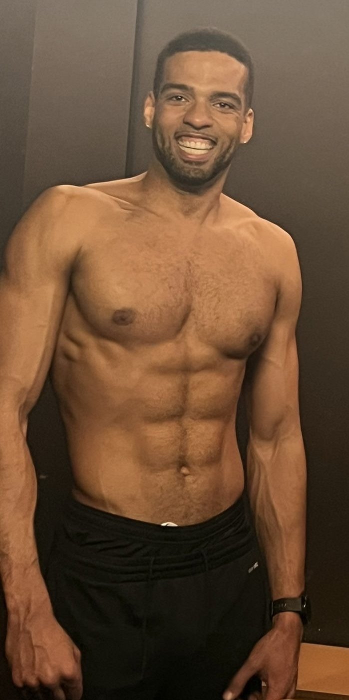 Manu today, smiling, defined torso against a dark background