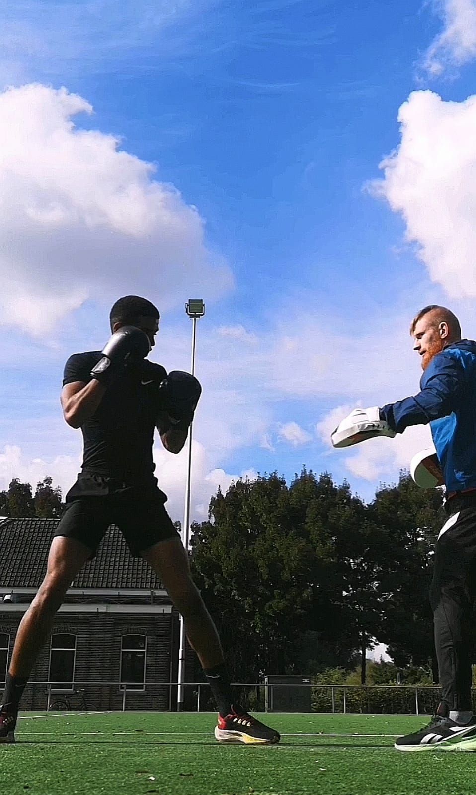 Manu boxing with gloves on, his coach holding the pads, on a grass field under a blue sky