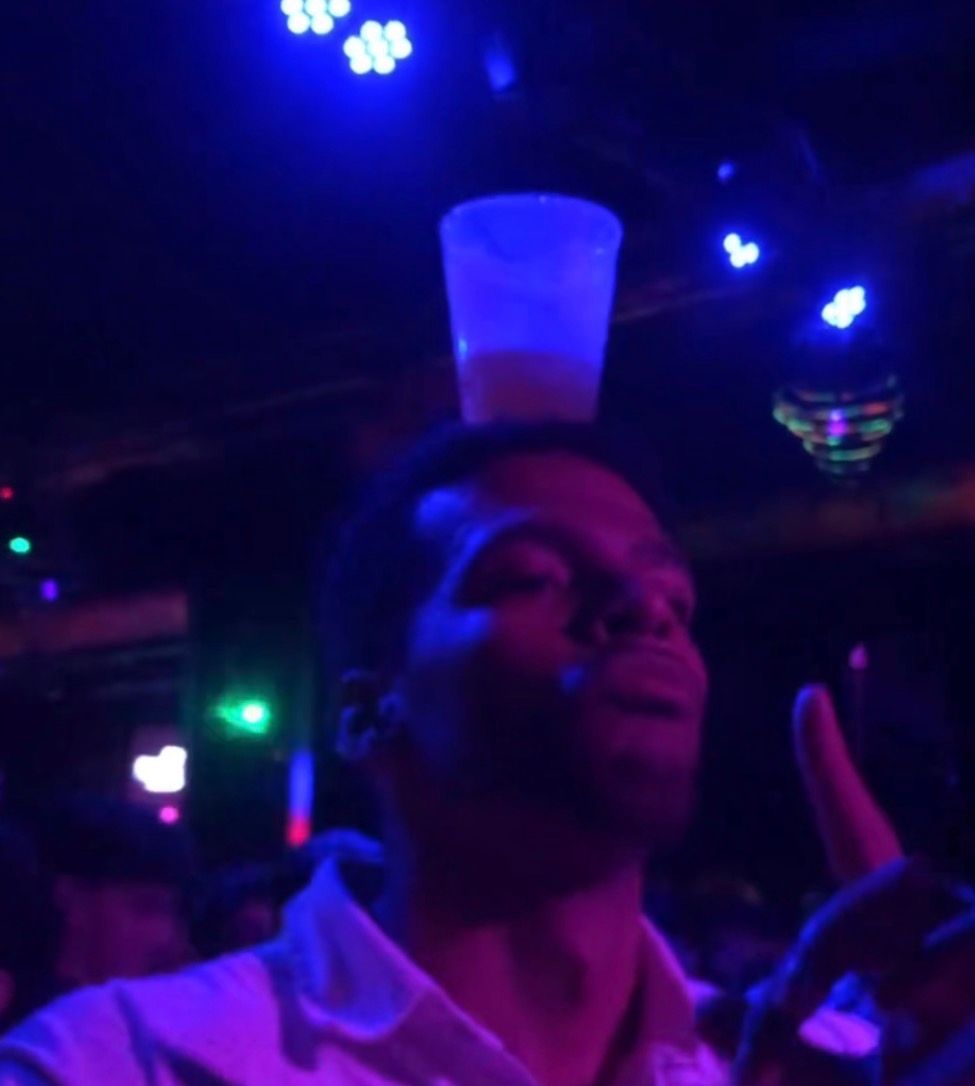 Manu at a club under blue lights, balancing a glass on his head